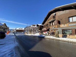 Una calle nevada en un pueblo, con edificios a ambos lados, incluyendo una tienda con un letrero que dice "SKIMIUM LOCATION".