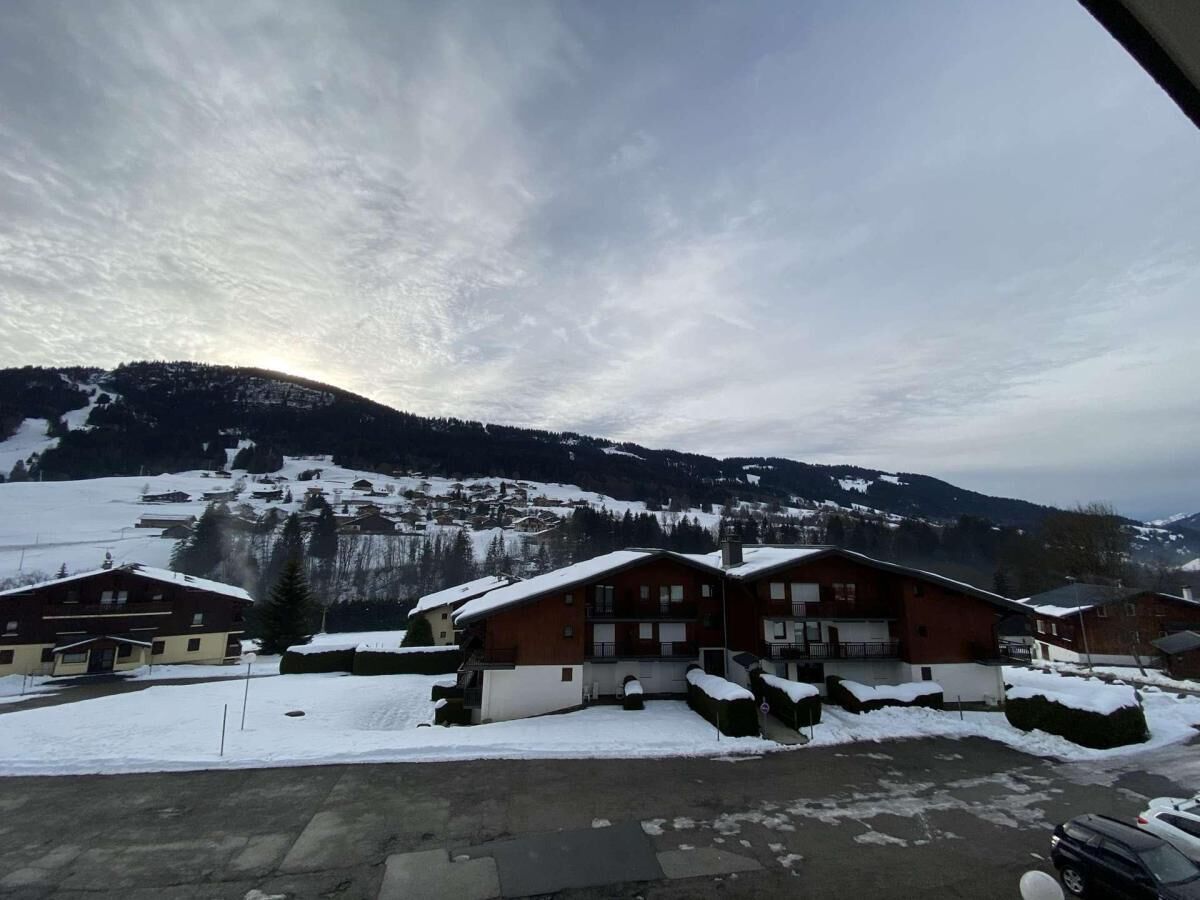 A snow-covered village with wooden buildings, a ski slope in the background, and a clear sky.