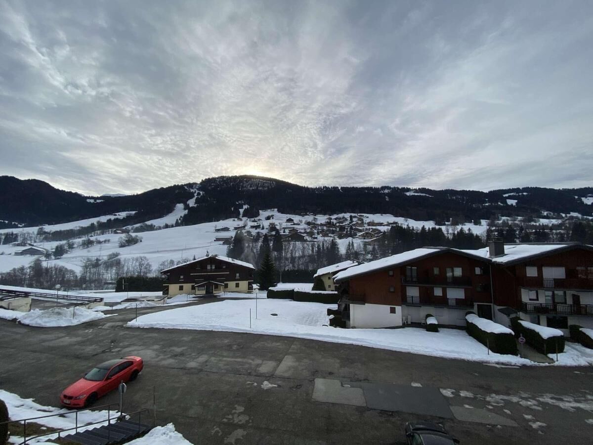 Uma aldeia coberta de neve, com edifícios, uma área de estacionamento e montanhas ao fundo.