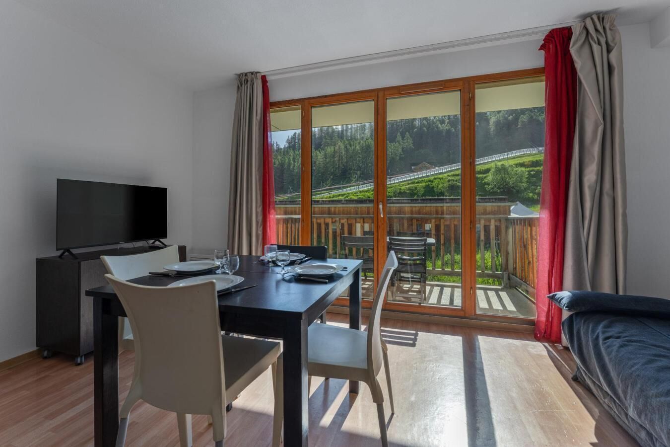Ein Zimmer mit Esstisch, Stühlen, Fernseher und einem Balkon mit Blick auf einen Wald und eine Brücke.