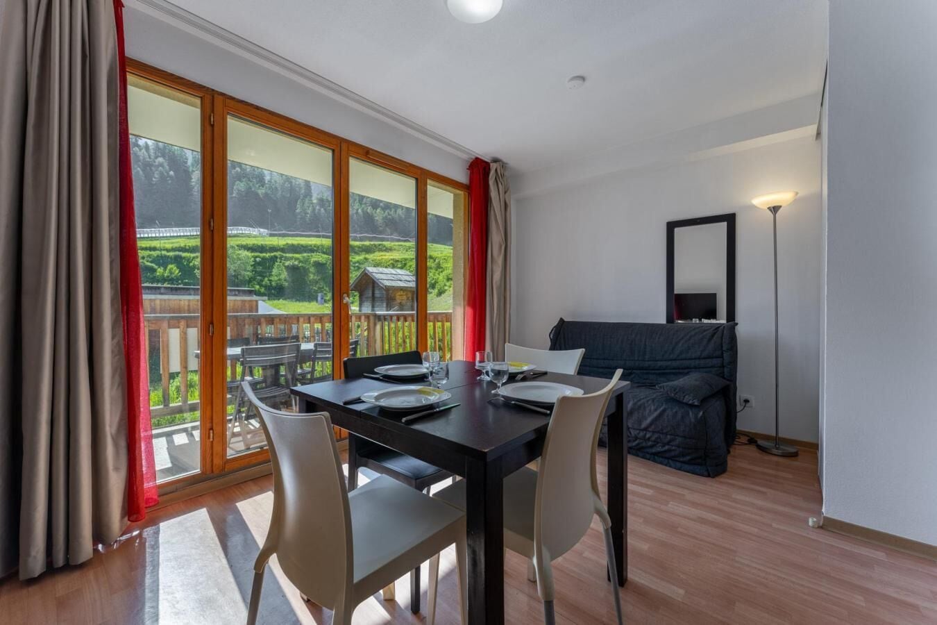 A dining area with a table set for two, a sofa, and a view of a green landscape through large windows.