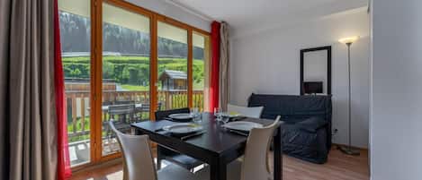 A dining area with a table set for two, a sofa, and a view of a green landscape through large windows.