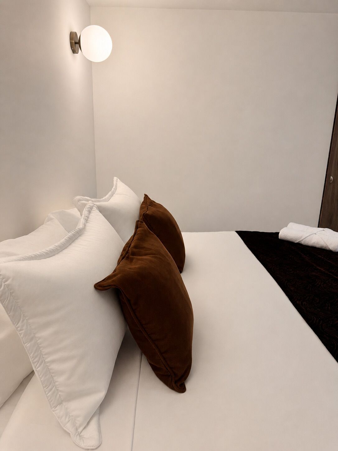 Una cama bien hecha con almohadas blancas y marrones, una manta oscura y una toalla blanca al lado.