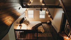 Un dormitorio estilo loft con techo inclinado, vigas de madera, una cama con cubrecama blanco, dos ventanas y un ventilador de techo.