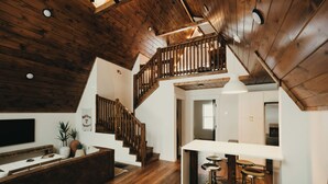 Un espacio habitable tipo loft con escalera de madera, una cocina con armarios blancos y una zona de bar con taburetes.