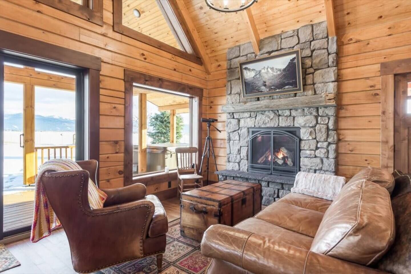 A log cabin interior with a stone fireplace, leather furniture, and a view through large windows.