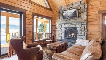 A log cabin interior with a stone fireplace, leather furniture, and a view through large windows.