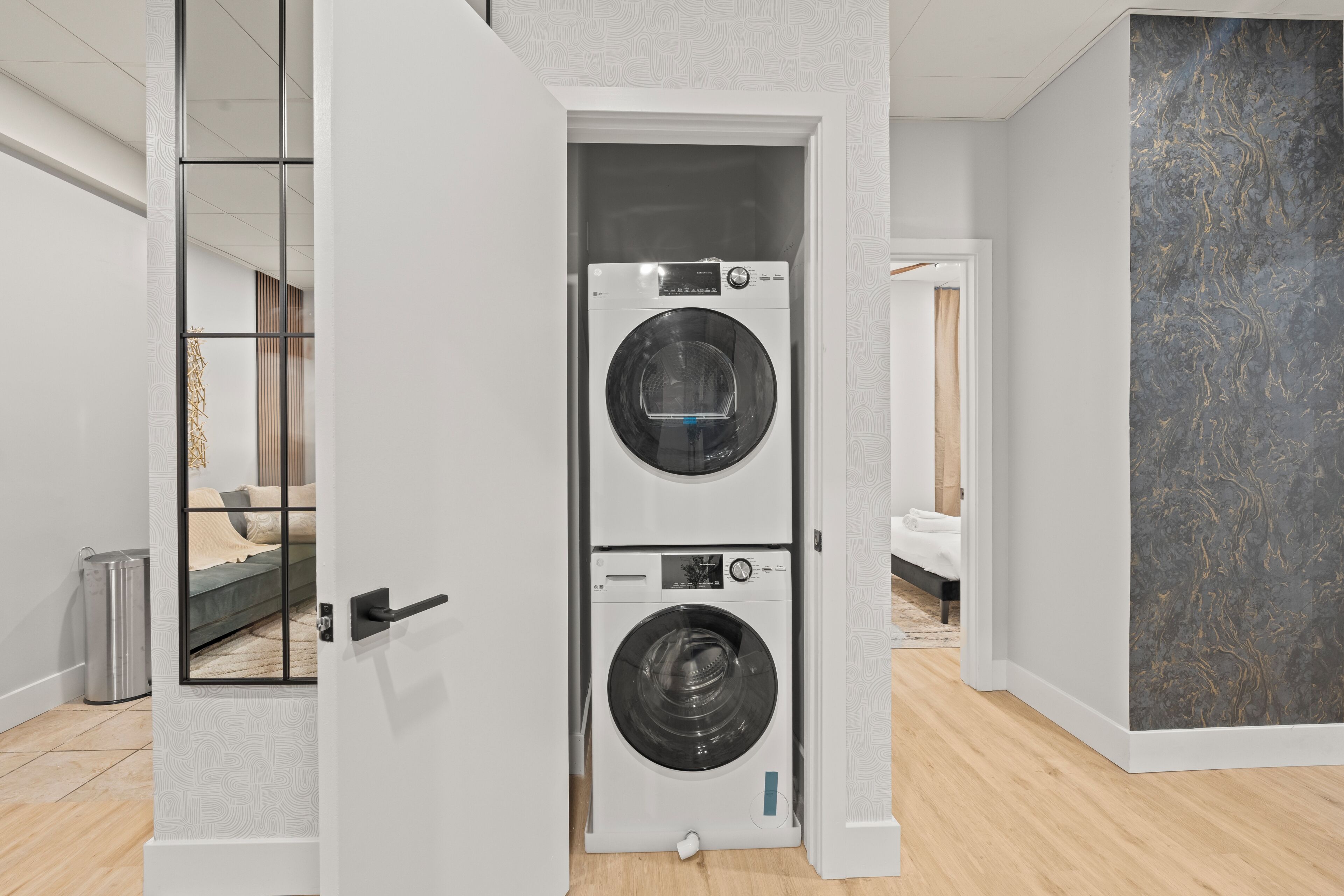 A modern laundry room with two stacked washing machines, a visible trash bin, and a bed in the background.