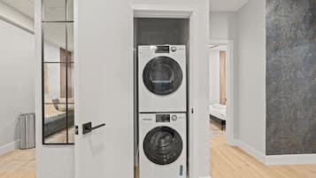 A modern laundry room with two stacked washing machines, a visible trash bin, and a bed in the background.