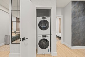A modern laundry room with two stacked washing machines, a visible trash bin, and a bed in the background.