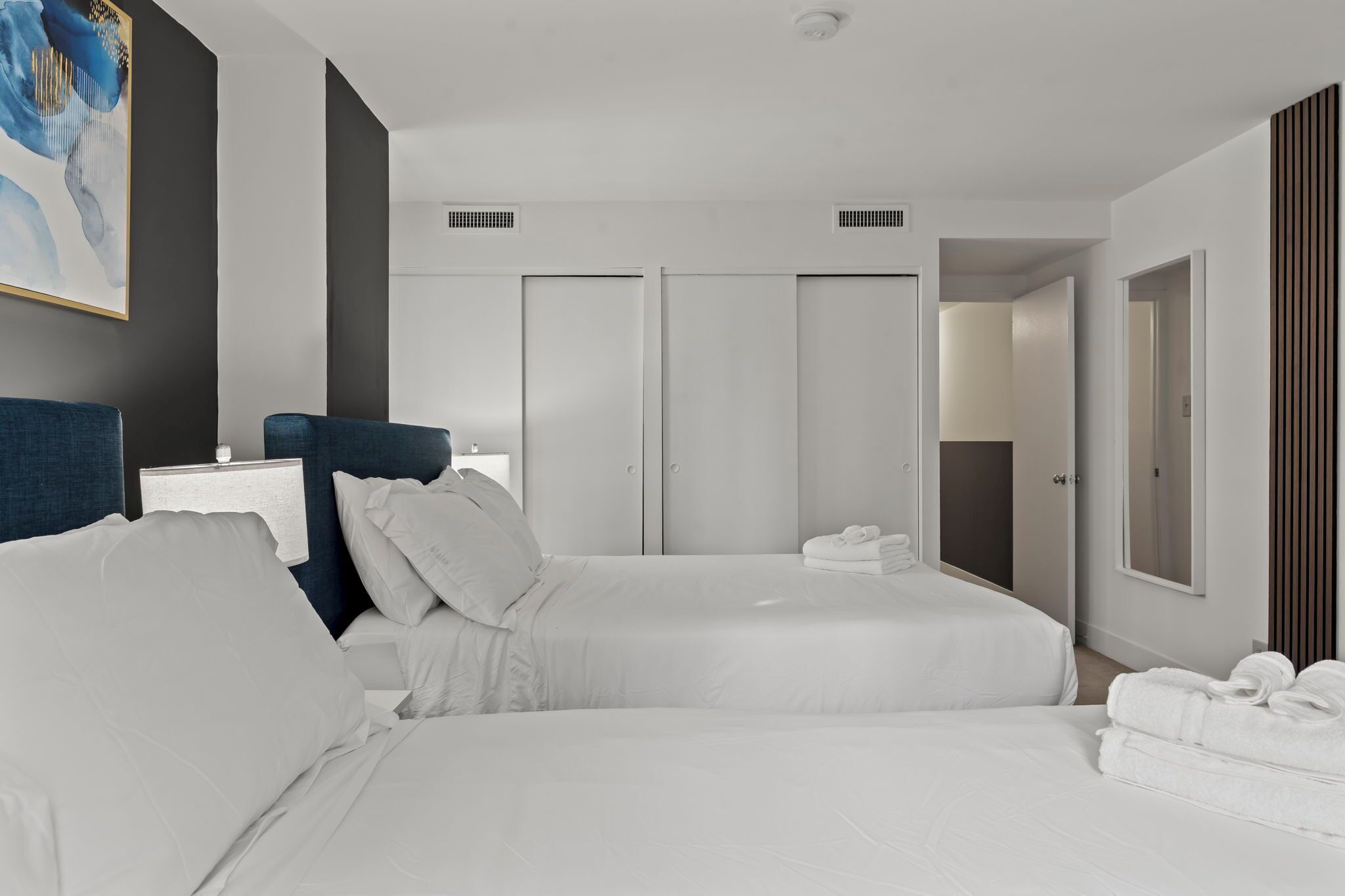 A modern hotel room with two beds, a large painting on the wall, and a view of the hallway.