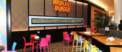 A restaurant interior with a checkered wall pattern, a neon sign reading 'MRBEAST BURGER', and multiple tables and chairs.
