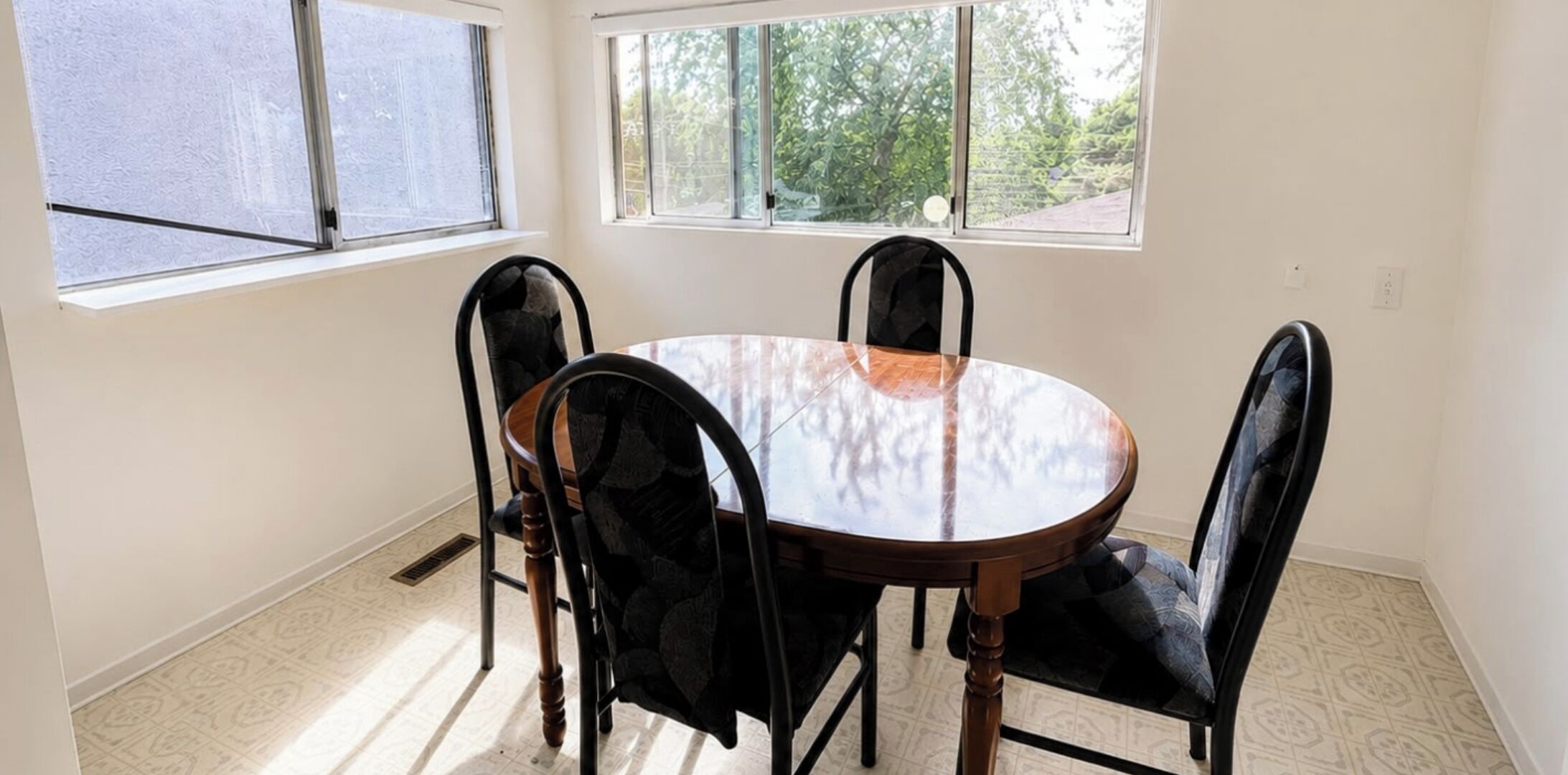 A dining area with a round table and four chairs, large windows, and a patterned floor.