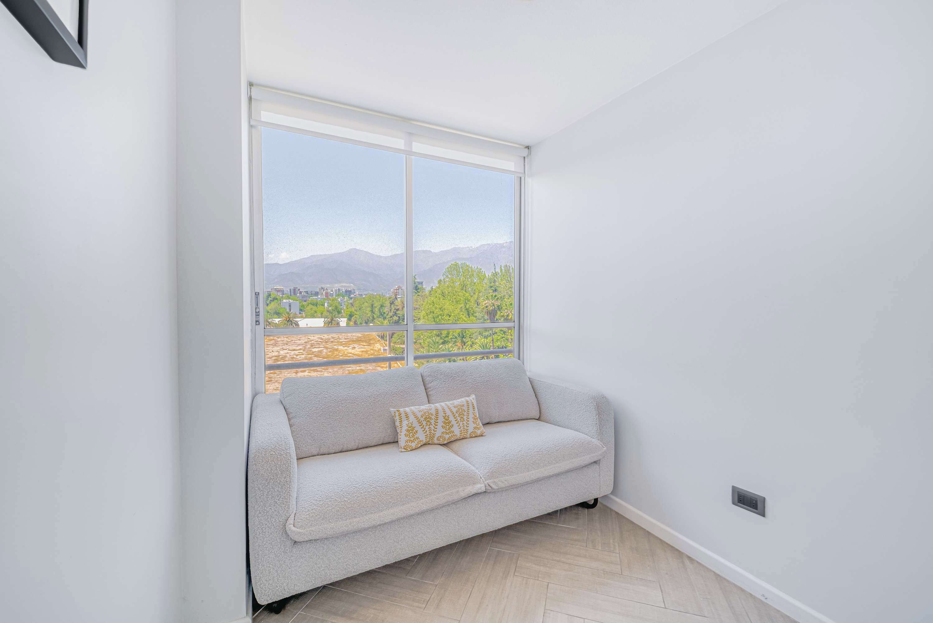 Quarto com um sofá de cor clara, uma almofada estampada e uma grande janela com vista para vegetação e montanhas.