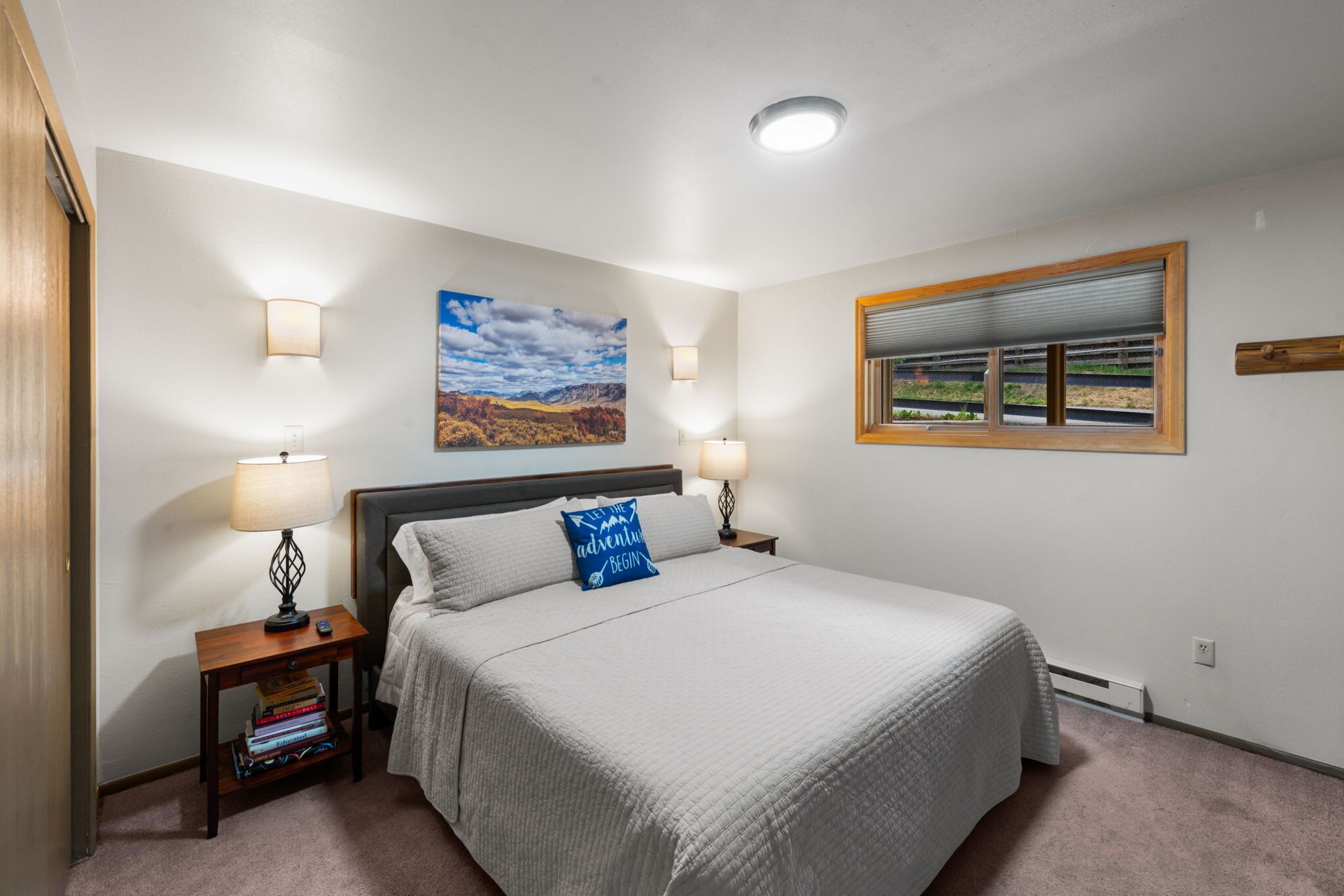 A bedroom with a bed, two bedside lamps, a nightstand with books, a painting, and a window with blinds.