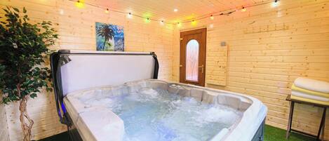 A hot tub in a room with wooden walls, a door, a small table with cushions, and a plant.