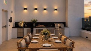 Uma área de refeições moderna com uma mesa de madeira posta para seis pessoas, uma cozinha com eletrodomésticos de aço inoxidável e vista para a cidade através de uma janela.