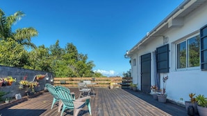 Une terrasse en bois avec des meubles d’extérieur, un bâtiment blanc doté de grandes fenêtres, et en arrière-plan, des arbres et un ciel dégagé.