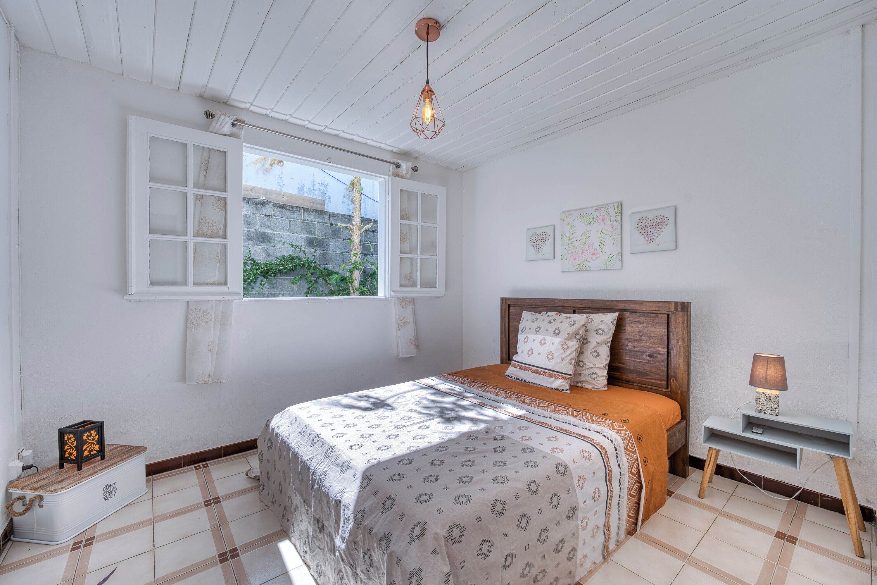 A bedroom with a bed, a bedside table, a hanging light fixture, a window, and a wall with two framed pictures.