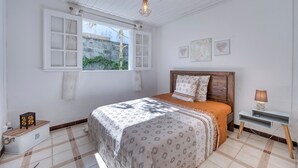 Une chambre à coucher comprenant un lit, une table de chevet, un luminaire suspendu, une fenêtre et un mur orné de deux tableaux encadrés.