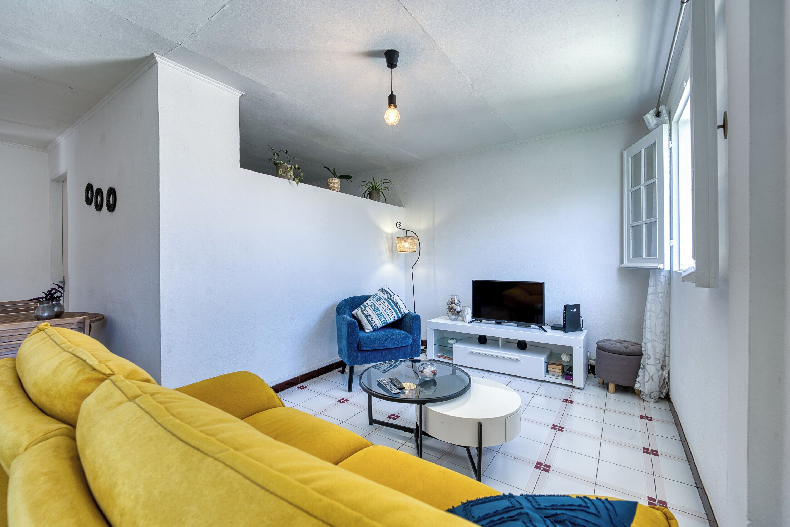 A living room with a yellow sofa, a blue armchair, a glass coffee table, a white entertainment unit, and a TV.