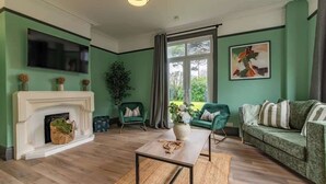 Ett vardagsrum med en öppen spis, gröna fåtöljer, en soffa, en platt-TV och ett stort fönster med utsikt över grönområden.