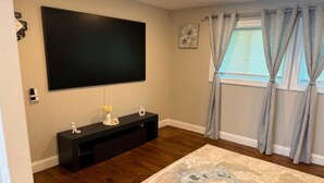 A room with a large flat-screen TV mounted on the wall, a dark wooden floor, a beige rug, and a window with curtains. - Cozy 1-Bedroom Apartment in Peaceful Commack with Air Conditioning & C. Monoxide (Commack)