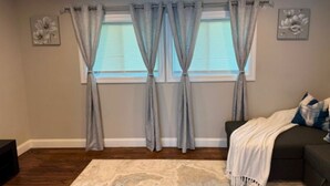 A living room with a dark-colored sofa, a patterned rug, and a window with grey curtains. - Cozy 1-Bedroom Apartment in Peaceful Commack with Air Conditioning & C. Monoxide (Commack)