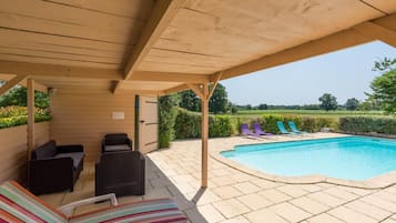 A covered outdoor area with a pool, lounge chairs, and a view of the countryside.