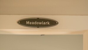 Et soveværelse med seng, et natbord, et vindue med persienner, og et skilt over sengen med teksten 'Meadowlark'.