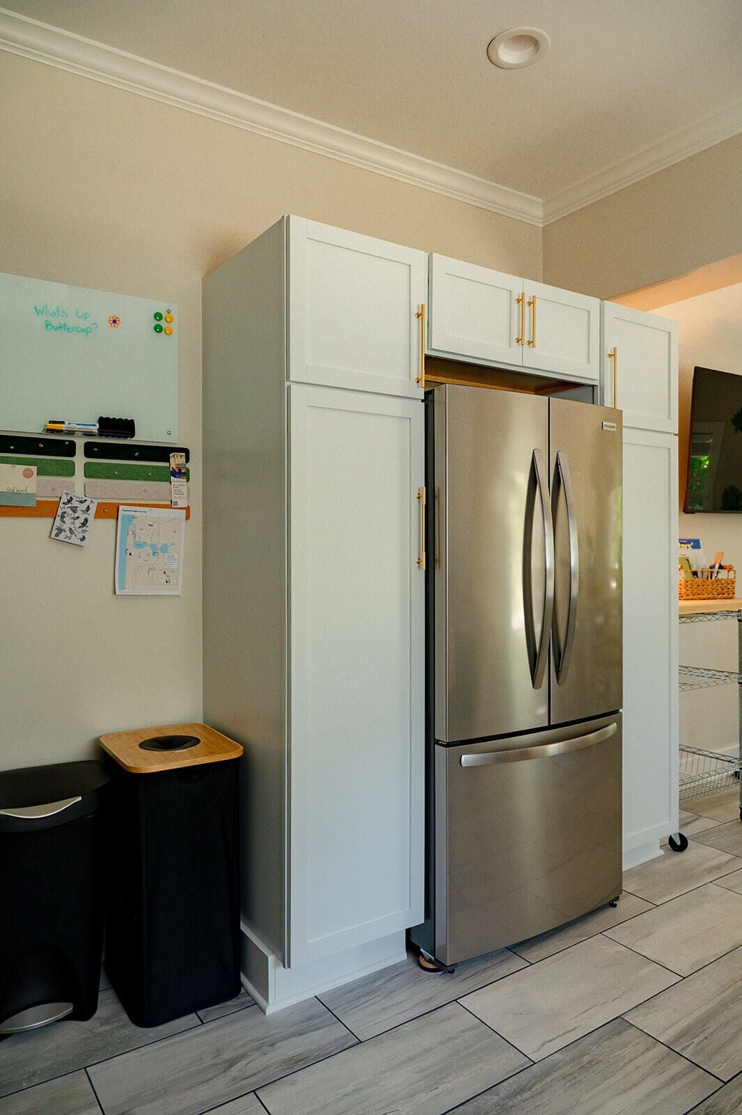 A kitchen with a stainless steel refrigerator, white cabinets, a black trash bin, and a whiteboard on the wall.