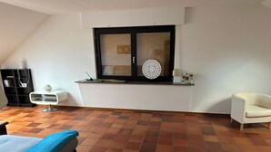 A room with a tiled floor, a bed, a white chair, a small table, a window with a decorative item, and a black bookshelf. - Find peace and discover yourself (Hürtgenwald)
