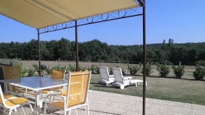 An outdoor patio with a table and chairs, a covered area, and a view of a grassy field and trees. - Petit Gite de la Mondette (LES-SALLES-DE-CASTILLON)