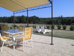 Een buitenterras met een tafel en stoelen, een overdekt gedeelte en uitzicht op een grasveld en bomen.