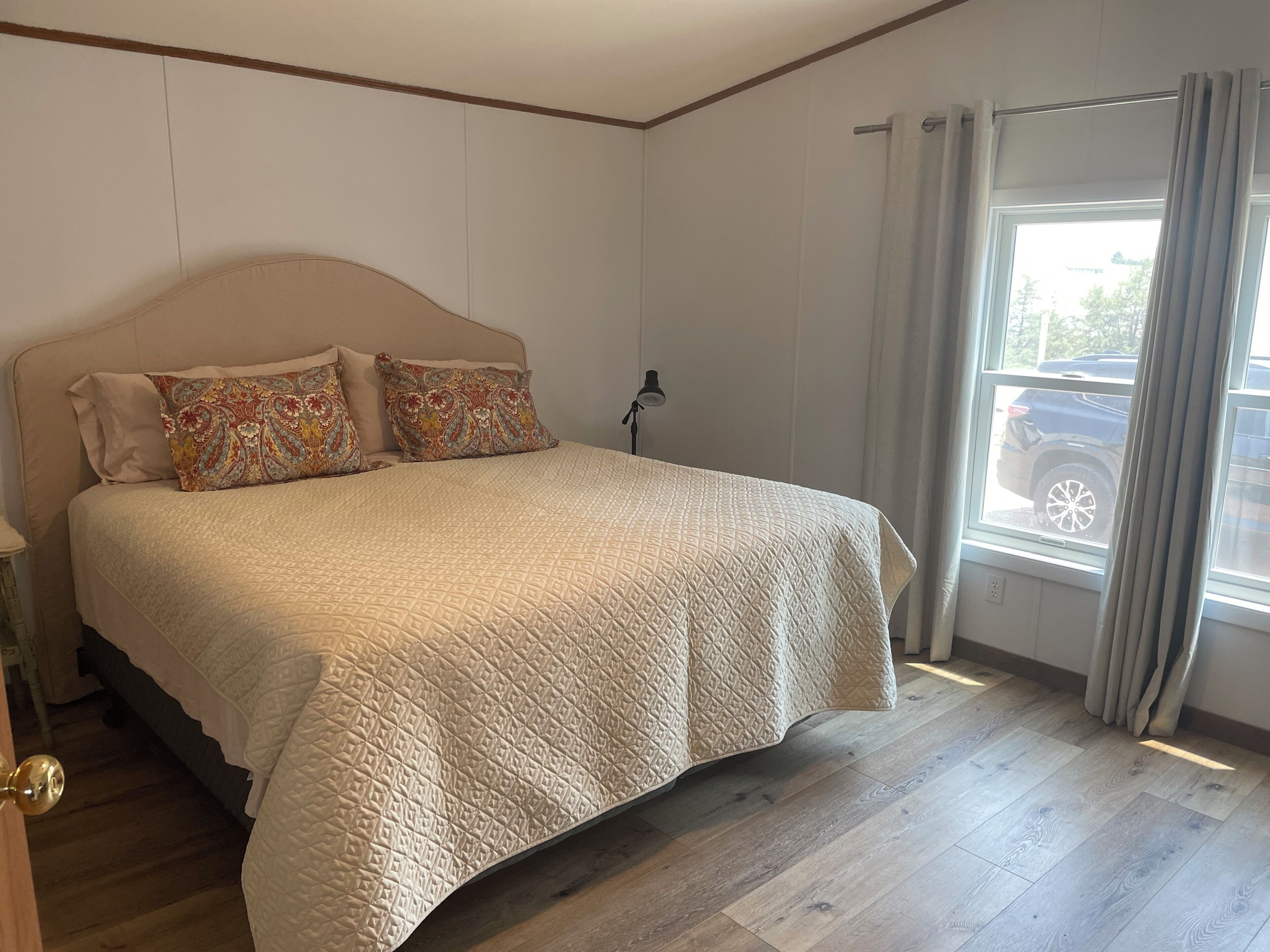 A bedroom with a bed, a nightstand, a lamp, a window with curtains, and a view of a parked car outside.