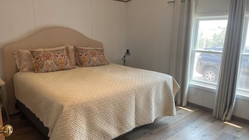 A bedroom with a bed, a nightstand, a lamp, a window with curtains, and a view of a parked car outside.