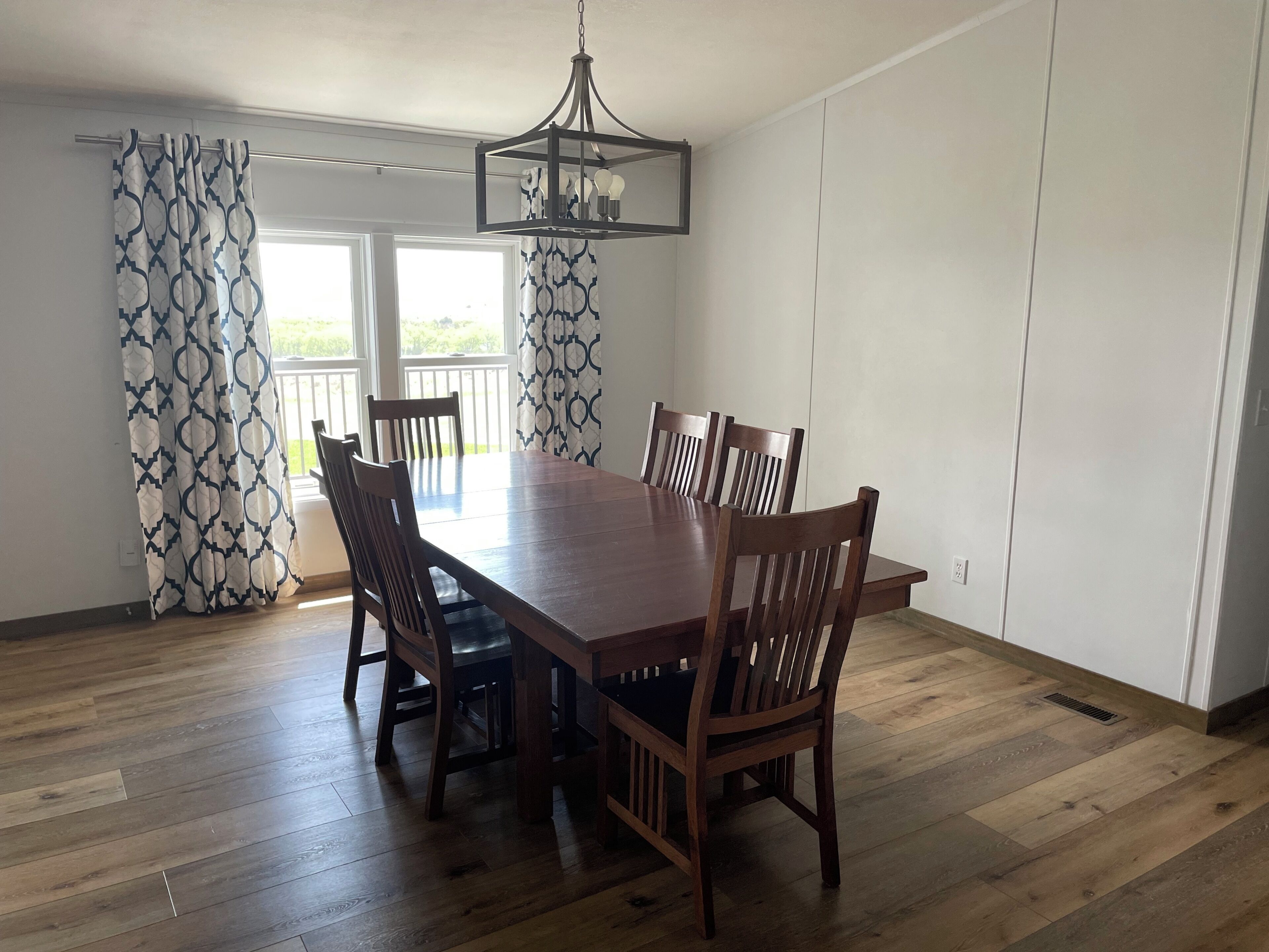 Un coin repas avec une table et des chaises en bois, un luminaire suspendu et une vue sur l’extérieur à travers une fenêtre ornée de rideaux à motifs.