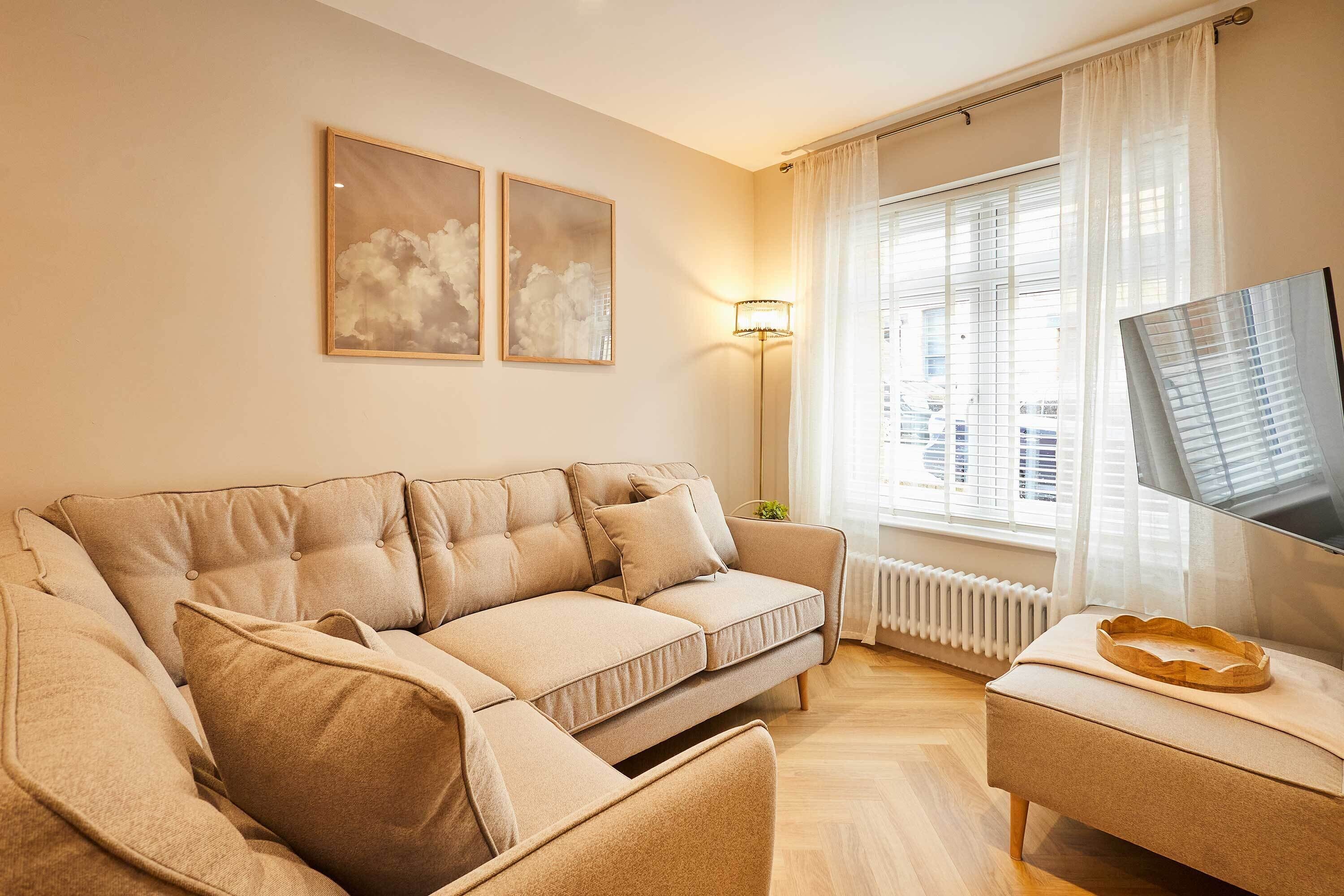 Una sala de estar con un sofá seccional color beige, un pequeño otoman, un televisor de pantalla plana y una ventana con cortinas traslúcidas.