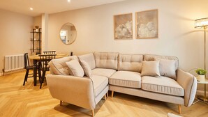 A living room with a beige sectional sofa, a round dining table with chairs, a small side table with a lamp, a radiator, and two framed cloud paintings on the wall. - Host Stay Belvedere Terrace (Scarborough)