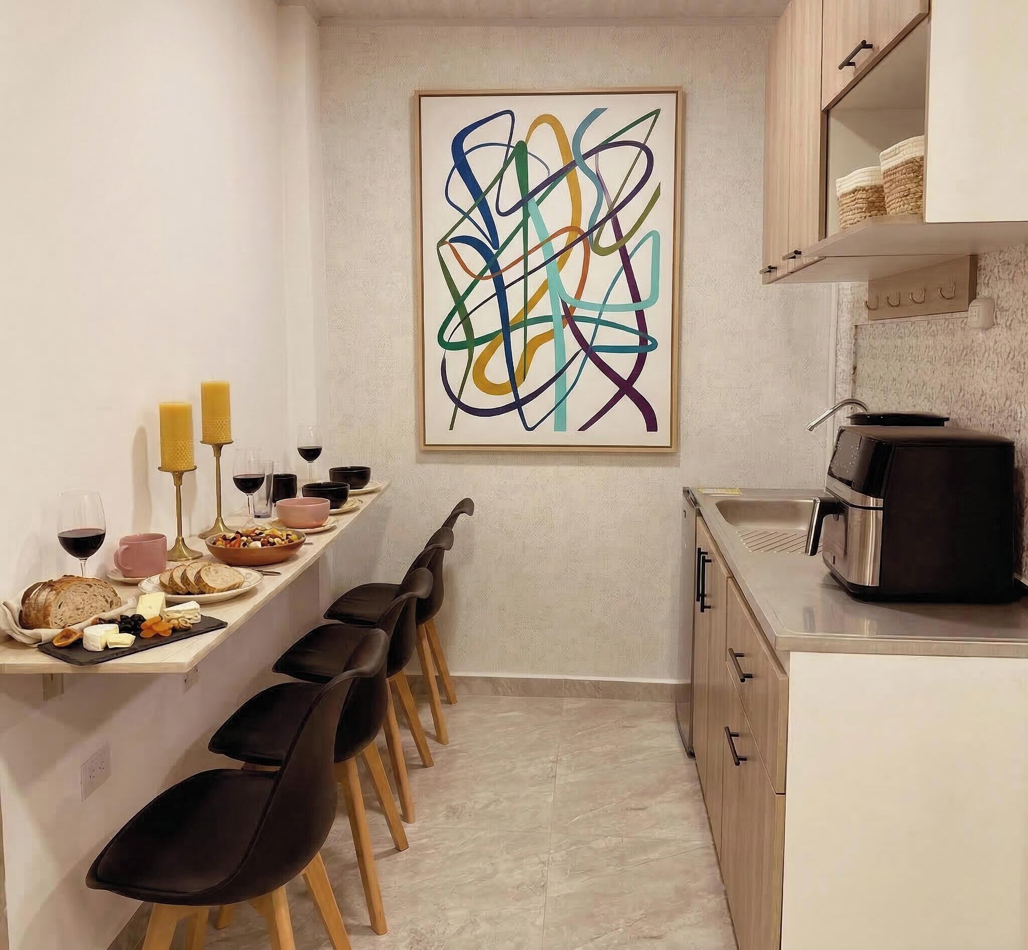 Une cuisine compacte avec un coin repas, agrémentée d’un tableau abstrait moderne, d’une machine à café et d’une variété d’aliments disposés sur le comptoir.