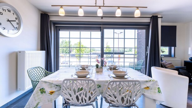 Een eetgedeelte met een tafel voor twee, een klok aan de muur en uitzicht door grote ramen.