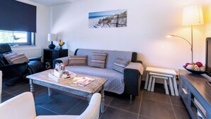 Een woonkamer met een bank, salontafel, lamp, schilderij aan de muur en een raam met zonwering.