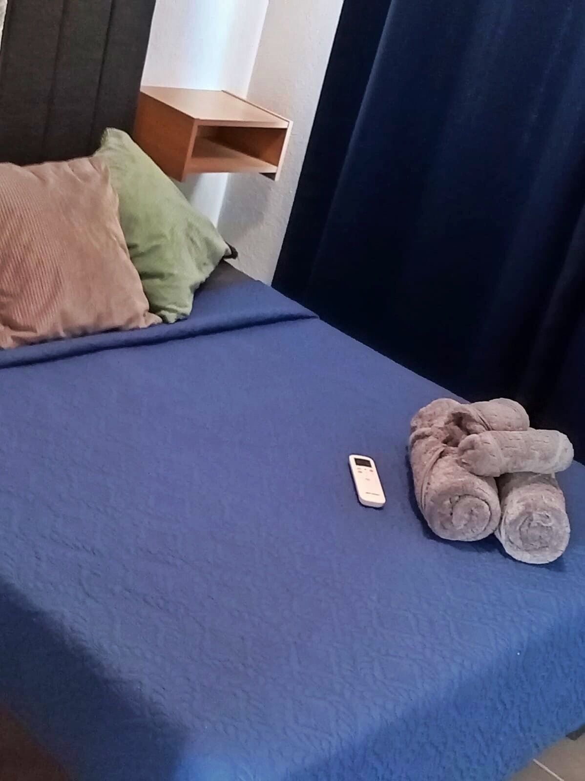 Ein Bett mit blauer Bettdecke, zwei Kissen, einem gefalteten Handtuch und einer weißen Fernbedienung.
