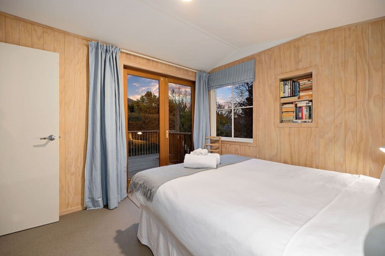 Un dormitorio con cama, una ventana con cortinas, una estantería y una puerta que da a un balcón.