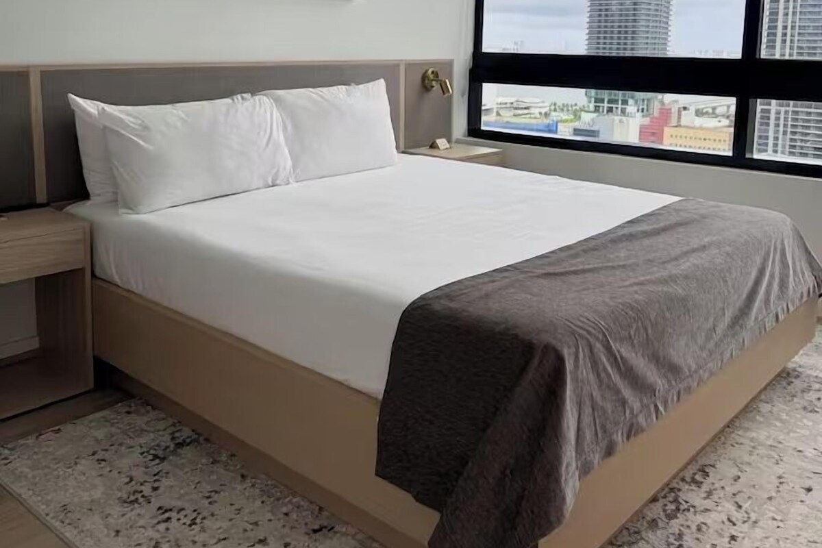 A hotel room with a large bed, a nightstand, a modern abstract painting, and a view of a tall building through a large window.