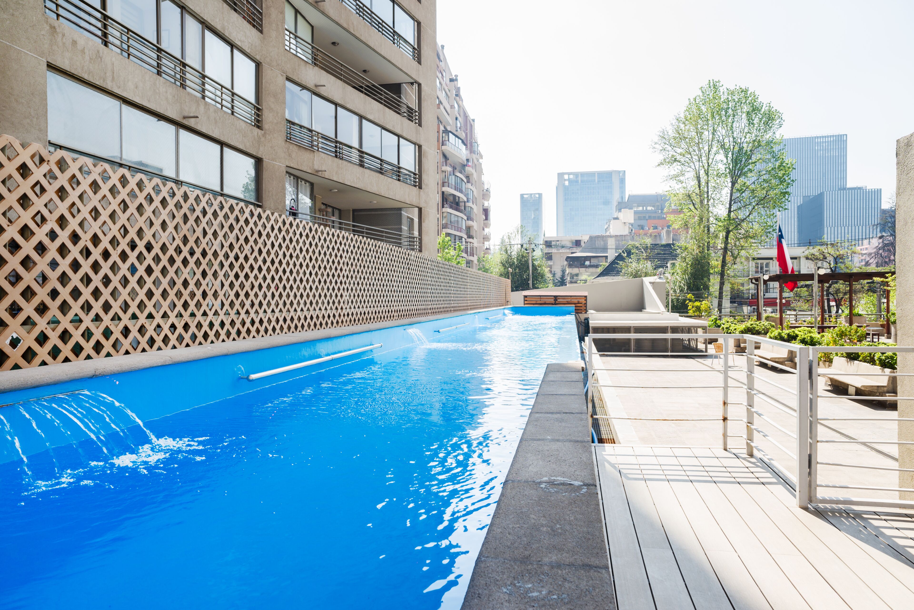 A swimming pool with a wooden deck, a lattice wall, and a cityscape in the background.