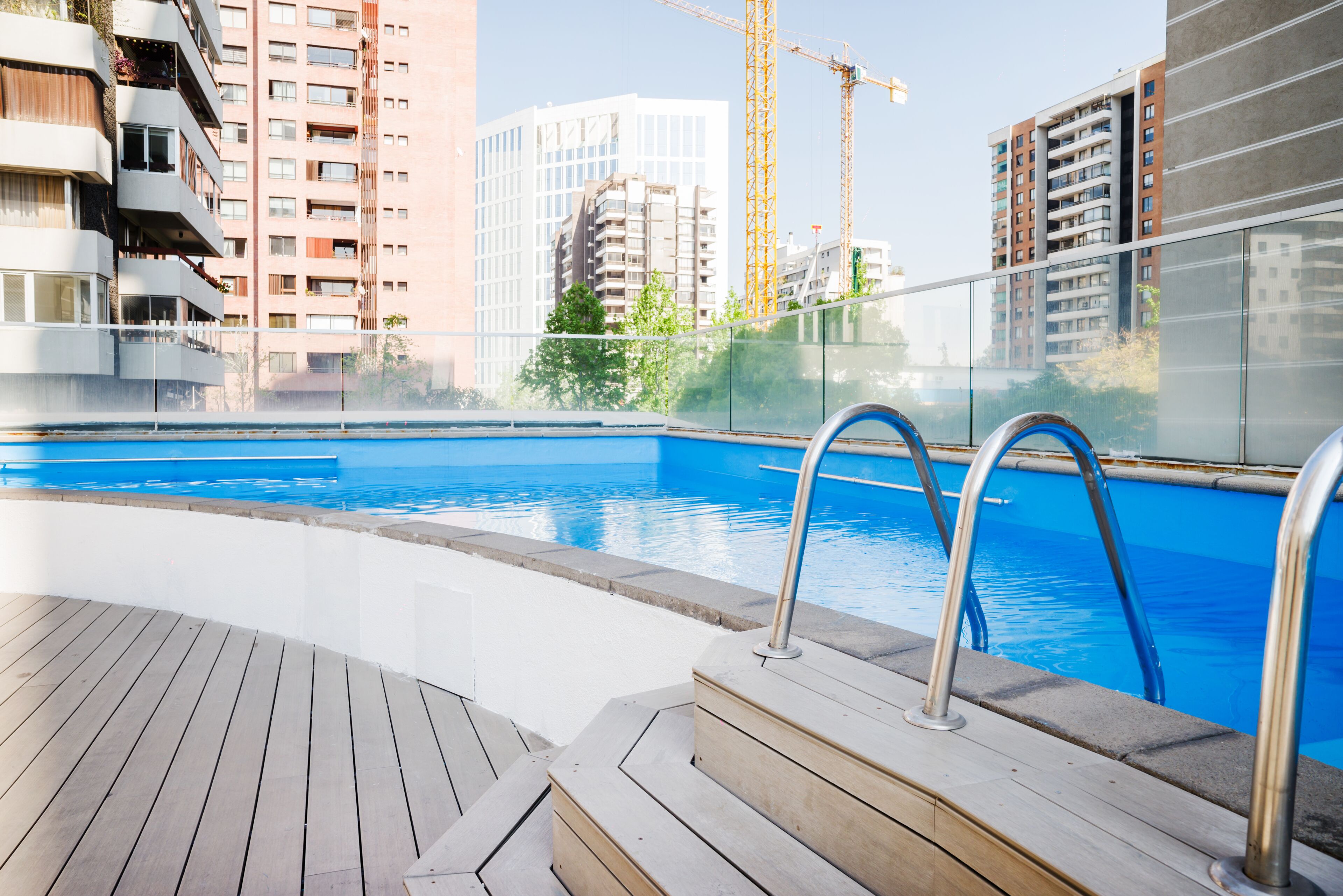 Una piscina con barandillas de metal, terraza de madera y edificios altos al fondo.