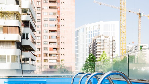 Ein Schwimmbecken mit Metallgeländern, einer Holzterrasse und modernen Gebäuden im Hintergrund.