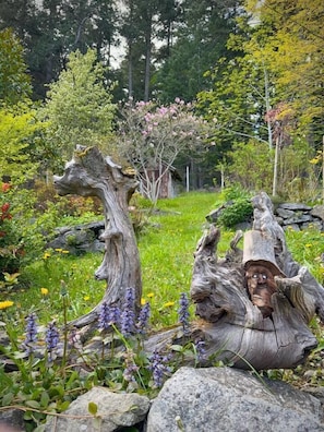 Un jardín con dos grandes tocones, uno con un rostro tallado, rodeado de rocas y flores moradas.
