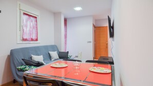 Ein kompakter Wohnraum mit Esstisch, Stühlen, einer Couch, einem an der Wand montierten Fernseher und einem Fenster mit Vorhängen.
